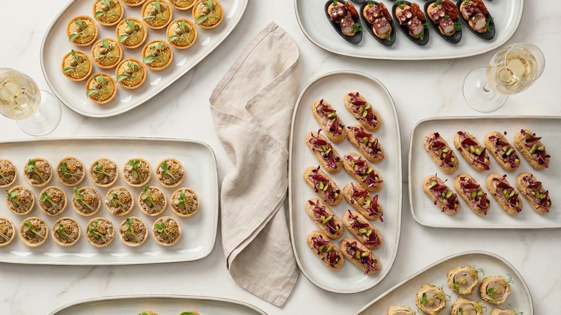 Assorted small canapés on white plates with a beige napkin and glasses of white wine on a light surface.